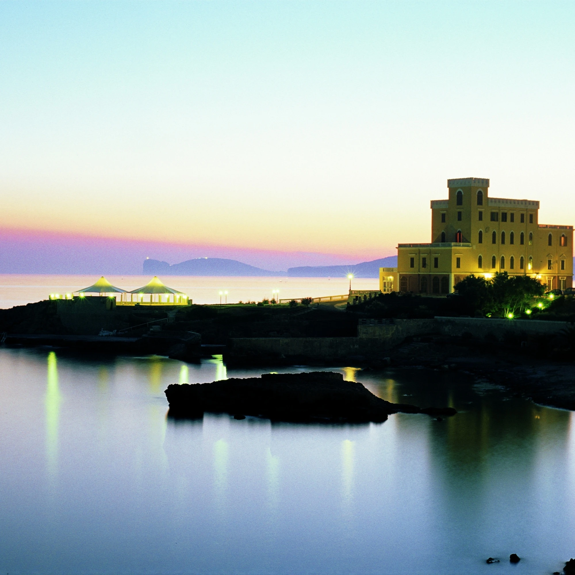 Veduta notturna di Villa Las Tronas Hotel & Spa illuminata sul mare di Alghero, con Capo Caccia all'orizzonte e riflessi sull'acqua.