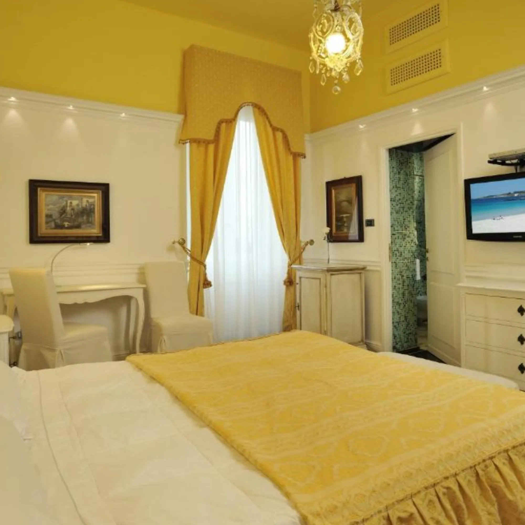 Angolo della camera matrimoniale di Villa Las Tronas Hotel & Spa, Alghero, con scrivania d'epoca, televisione e porta aperta sul bagno in mosaico azzurro.