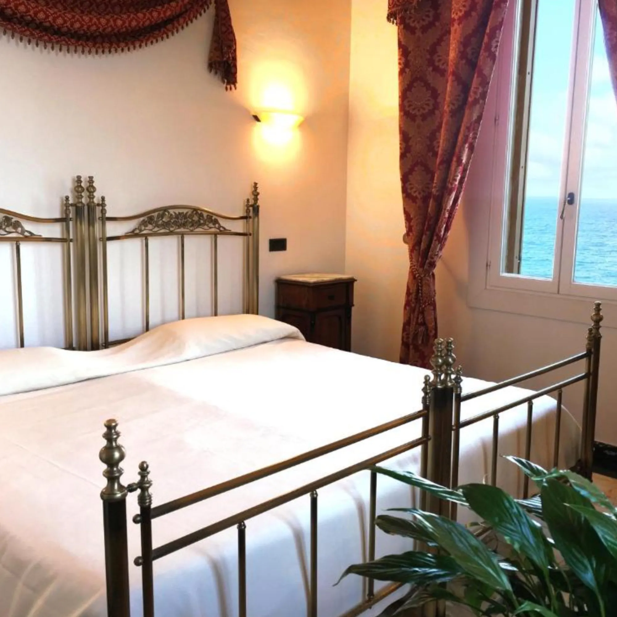 Particolare di una camera matrimoniale presso Villa Las Tronas Hotel & Spa, con letto in ottone lavorato, tende classiche e vista sul mare di Alghero.