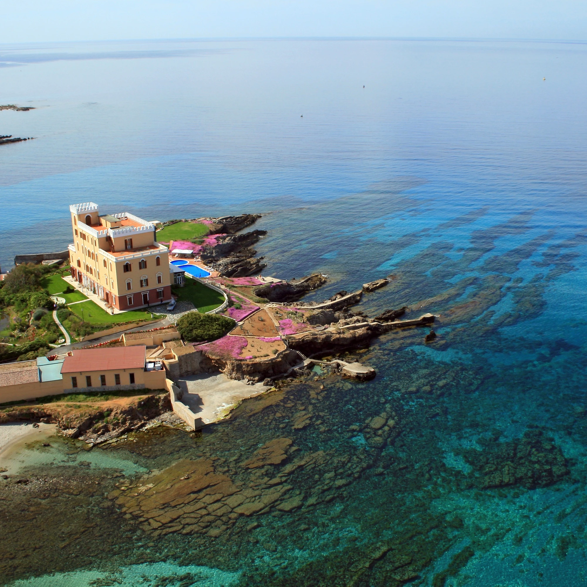 Veduta aerea di Villa Las Tronas Hotel & Spa ad Alghero, circondata dal mare cristallino della Sardegna e scogliere dorate.